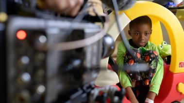 Little boy in hospital in toy car playing with cameraman. Project sunshine behind the scenes film shoot. 5-part promotional campaign to highlight the scope and impact of their work. Pediatric patients. Non profit. Production services.